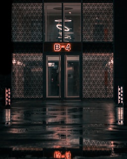 A building entrance with a double door and a glowing sign displaying 'B-4' above it. The exterior walls feature decorative patterns, and an artistic light fixture with circular designs is visible through the upper windows. The wet ground reflects the scene, enhancing the moody ambiance.