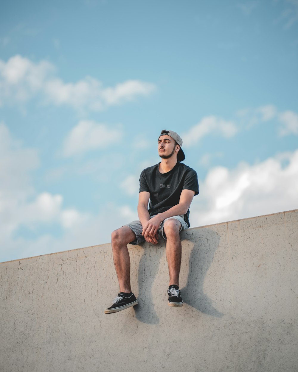 Mann in schwarzem Rundhals-T-Shirt sitzt auf grauer Betonwand unter blauem Himmel während