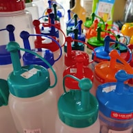 A collection of colorful plastic bottles with lids and pour spouts, ranging in colors including blue, red, green, and orange. Each bottle has a price sticker attached, and some have a clear body. They are neatly arranged on a shelf in a store setting.