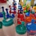 A collection of colorful plastic bottles with lids and pour spouts, ranging in colors including blue, red, green, and orange. Each bottle has a price sticker attached, and some have a clear body. They are neatly arranged on a shelf in a store setting.