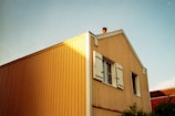 A wooden house with yellow siding and white trim is bathed in sunlight. It features a chimney protruding from the roof, with one window on the side and another with open shutters. The sky is clear and blue, and part of another building with red roofing is visible on the right.