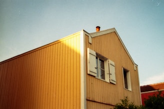 A wooden house with yellow siding and white trim is bathed in sunlight. It features a chimney protruding from the roof, with one window on the side and another with open shutters. The sky is clear and blue, and part of another building with red roofing is visible on the right.