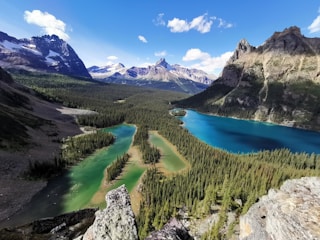 A stunning mountain landscape featuring two lakes of different colors, green and blue, surrounded by dense forests. Snow-capped mountains and rocky cliffs dominate the background under a bright blue sky with scattered clouds.