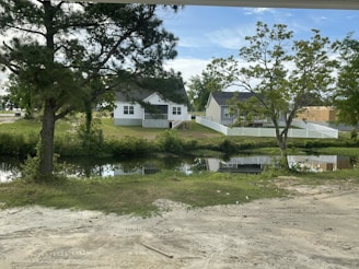 Two white houses with pitched roofs and front porches are situated behind a small body of water, surrounded by greenery and trees. A white picket fence outlines the property. The sky is clear with some clouds, and the reflection of the houses and sky can be seen in the water.