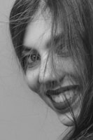 A close-up black and white portrait of a person with long hair partially covering their face. The focus is on the person's eyes and smile, conveying a sense of joy and spontaneity. The hair is slightly messy, adding a natural and candid feel to the image.