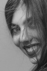 A close-up black and white portrait of a person with long hair partially covering their face. The focus is on the person's eyes and smile, conveying a sense of joy and spontaneity. The hair is slightly messy, adding a natural and candid feel to the image.
