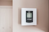 A digital thermostat is mounted on a beige wall. The thermostat has an LED screen displaying the temperature settings and controls below it. The overall look is clean and minimalistic.