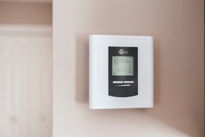 A digital thermostat is mounted on a beige wall. The thermostat has an LED screen displaying the temperature settings and controls below it. The overall look is clean and minimalistic.