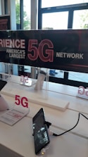 A display inside a store features signage promoting America's largest 5G network. Two smartphones are mounted on stands, one with a visible price tag offering a Samsung Galaxy A32 for $504.00. The background shows clear glass doors and windows, providing natural light to the interior.