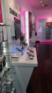 A retail store interior displaying modern electronic devices on white shelves. The walls are accented with pink lighting, and products from Samsung are prominently featured. In the foreground, several smartphones are showcased on stands, with various accessories visible on racks. The space is sleek and well-organized, emphasizing a contemporary technology theme.