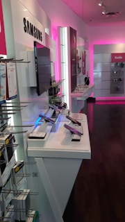 A retail store interior displaying modern electronic devices on white shelves. The walls are accented with pink lighting, and products from Samsung are prominently featured. In the foreground, several smartphones are showcased on stands, with various accessories visible on racks. The space is sleek and well-organized, emphasizing a contemporary technology theme.