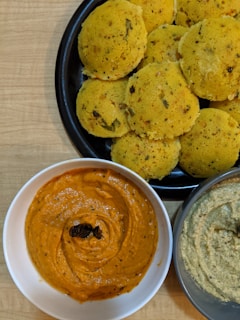 A plate of round, yellowish savory cakes with herbs served alongside two bowls of chutney. One bowl contains a vibrant orange chutney with a garnish on top, and the other has a beige chutney with visible seasoning.