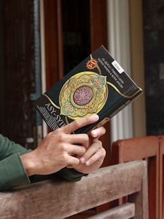 A person holding a book with a decorative cover featuring Arabic script and intricate patterns. The scene is set against a wooden background, suggesting calmness and introspection.
