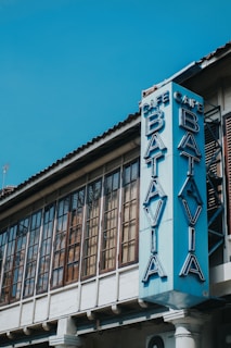 A vintage-style building with large, rectangular windows featuring a vertical sign reading 'Cafe Batavia' in blue neon letters. The architectural design includes a tiled roof and wooden window frames.