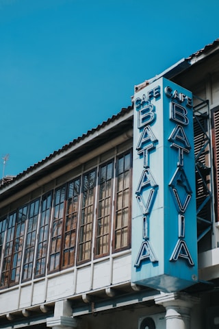 A vintage-style building with large, rectangular windows featuring a vertical sign reading 'Cafe Batavia' in blue neon letters. The architectural design includes a tiled roof and wooden window frames.