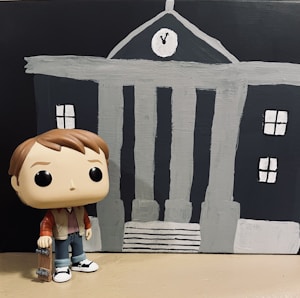 A toy figure resembles a young man wearing a red jacket and blue jeans, holding a skateboard. It stands in front of a painted backdrop depicting a stylized gray and black building with a clock showing a specific time. The building features several windows and pillars.