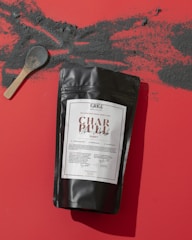 A black packet labeled 'Char Black Mask Purify' is placed on a red surface. A wooden spoon with black powder rests nearby, surrounded by scattered black powder across the surface.