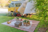A serene outdoor picnic setup with a yellow van in the background. The picnic area includes a large blanket, wicker baskets, flowers, and a red lantern. The scene is shaded by flowing white fabric from the van, and there is greenery surrounding the setting, including bushes on the right side.