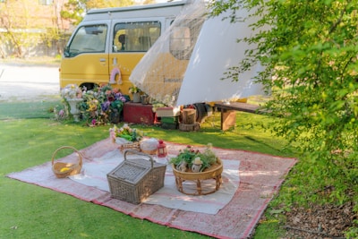 A serene outdoor picnic setup with a yellow van in the background. The picnic area includes a large blanket, wicker baskets, flowers, and a red lantern. The scene is shaded by flowing white fabric from the van, and there is greenery surrounding the setting, including bushes on the right side.