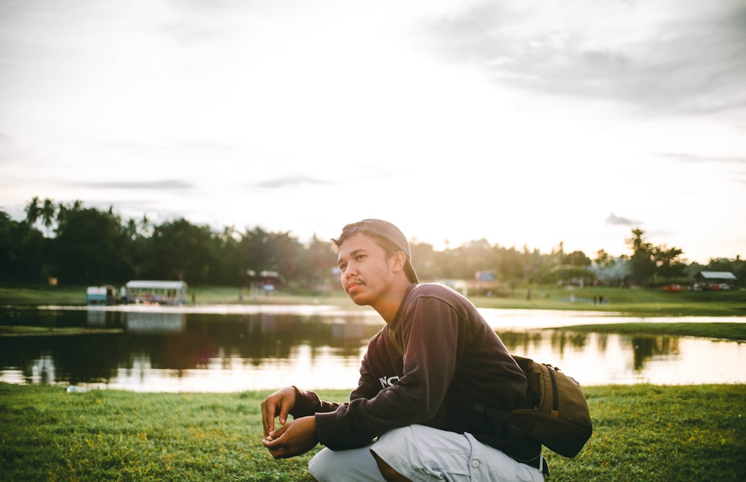 man in gray hoodie sitting on grass field near lake during daytime