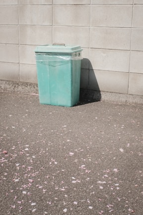 A mint green trash bin is positioned against a beige stone wall. The pavement in front of the bin is scattered with pink flower petals, suggesting a recent presence of flowers.