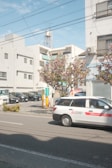 A street scene featuring a white car with 'Mitsubishi Electric' branding parked on a road. In the background are multiple parked vehicles in a lot, several modern buildings with balconies and windows, and a utility pole with wires overhead. The sky is partly cloudy with a soft blue hue. There is a tree with pink blossoms near the sidewalk, adding vibrancy to the urban setting.
