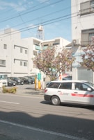 A street scene featuring a white car with 'Mitsubishi Electric' branding parked on a road. In the background are multiple parked vehicles in a lot, several modern buildings with balconies and windows, and a utility pole with wires overhead. The sky is partly cloudy with a soft blue hue. There is a tree with pink blossoms near the sidewalk, adding vibrancy to the urban setting.
