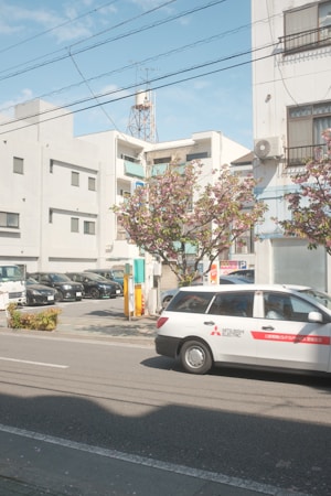 A street scene featuring a white car with 'Mitsubishi Electric' branding parked on a road. In the background are multiple parked vehicles in a lot, several modern buildings with balconies and windows, and a utility pole with wires overhead. The sky is partly cloudy with a soft blue hue. There is a tree with pink blossoms near the sidewalk, adding vibrancy to the urban setting.
