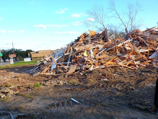 A large pile of debris consisting of wooden planks and broken materials is situated on a rough dirt ground. Sparse trees without leaves appear in the background along with a building and some road signs.