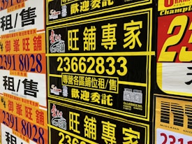 A collection of colorful advertisement posters displayed on a wall, featuring large, bold text in various fonts, primarily in Chinese characters, with some English text. Yellow, black, and red are the prominent color schemes used in the posters.