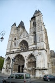 A historic stone church with two prominent towers featuring Gothic architectural elements like pointed arches, intricate carvings, and a large circular stained glass window above the entrance. A couple of modern cars are parked in front of it, and a lamp post is visible to the left.