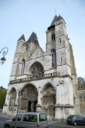 A historic stone church with two prominent towers featuring Gothic architectural elements like pointed arches, intricate carvings, and a large circular stained glass window above the entrance. A couple of modern cars are parked in front of it, and a lamp post is visible to the left.