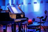 A grand piano with open sheet music on the stand is placed on a stage. The environment is dimly lit with a blue hue, providing a serene and focused ambiance. Surrounding the piano, there are chairs and musical stands, indicating a setup for a musical performance or rehearsal.