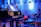 A grand piano with open sheet music on the stand is placed on a stage. The environment is dimly lit with a blue hue, providing a serene and focused ambiance. Surrounding the piano, there are chairs and musical stands, indicating a setup for a musical performance or rehearsal.