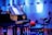 A grand piano with open sheet music on the stand is placed on a stage. The environment is dimly lit with a blue hue, providing a serene and focused ambiance. Surrounding the piano, there are chairs and musical stands, indicating a setup for a musical performance or rehearsal.