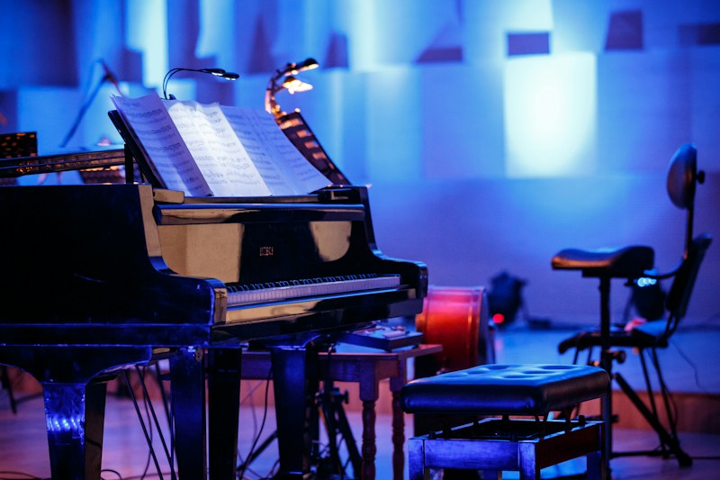 A grand piano with open sheet music on the stand is placed on a stage. The environment is dimly lit with a blue hue, providing a serene and focused ambiance. Surrounding the piano, there are chairs and musical stands, indicating a setup for a musical performance or rehearsal.