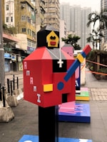 An abstract, colorful statue resembling a blocky humanoid figure holding a guitar is positioned in an urban environment. The figure features a red body, yellow face, and black head, with a white cross pendant around its neck. Surrounding the statue are tall buildings and a few palm trees, indicating a cityscape setting.