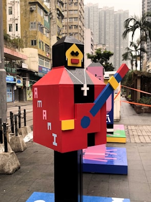 An abstract, colorful statue resembling a blocky humanoid figure holding a guitar is positioned in an urban environment. The figure features a red body, yellow face, and black head, with a white cross pendant around its neck. Surrounding the statue are tall buildings and a few palm trees, indicating a cityscape setting.