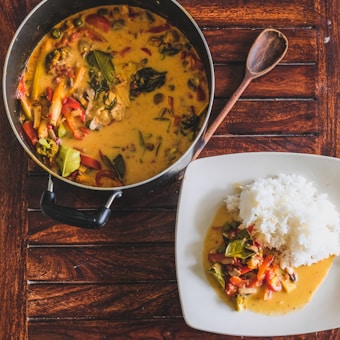 A pot filled with creamy curry containing assorted vegetables such as broccoli, red peppers, and leafy greens is placed on a wooden table. Next to the pot, there is a white square plate holding a serving of steamed white rice with a portion of the curry. A wooden spoon lies beside the pot.