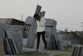 A person is standing among stacks of large, rectangular stone slabs arranged in an outdoor setting. The individual is wearing a white shirt and black pants, with one hand positioned under their chin as they appear to be in thought. In the background, there are power lines and a sparse amount of greenery.