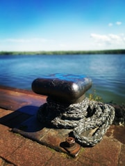 A close-up view of a metal bollard on a dock with a thick rope coiled around it. In the background, a body of water stretches out under a clear blue sky. The dock and bollard appear weathered, indicating frequent use.