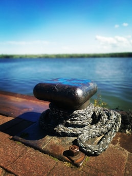 A close-up view of a metal bollard on a dock with a thick rope coiled around it. In the background, a body of water stretches out under a clear blue sky. The dock and bollard appear weathered, indicating frequent use.