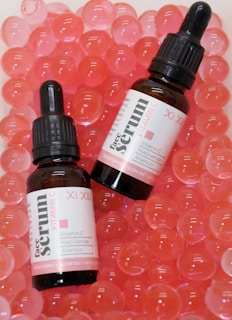 Two bottles of face serum with vitamin C are positioned on a surface filled with pink translucent gel-like beads. The bottles have black droppers and white labels with red and black text.