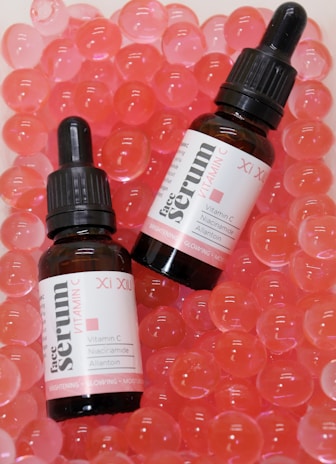 Two bottles of face serum with vitamin C are positioned on a surface filled with pink translucent gel-like beads. The bottles have black droppers and white labels with red and black text.
