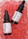 Two bottles of face serum with vitamin C are positioned on a surface filled with pink translucent gel-like beads. The bottles have black droppers and white labels with red and black text.