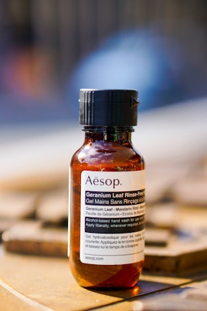 A small amber bottle labeled 'Aesop' containing a rinse-free hand wash. The label includes ingredients such as Geranium Leaf and Mandarin Rind. The bottle is placed on a flat surface with a blurred background, suggesting an outdoor or well-lit setting.
