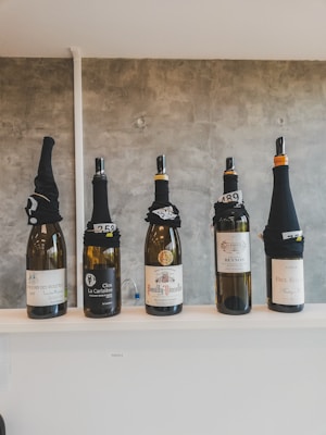 Five bottles of wine are displayed on a white shelf. Each bottle is covered at the top with a black fabric, concealing the cork and upper label, and is marked with white numbers indicating a possible blind tasting setup. The background is a plain gray wall.