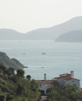 A peaceful coastal scene with a large villa in the foreground, surrounded by lush greenery. In the background, the calm sea stretches out towards a range of gently sloping hills. Several small boats are scattered across the water, contributing to the serene atmosphere.