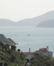A peaceful coastal scene with a large villa in the foreground, surrounded by lush greenery. In the background, the calm sea stretches out towards a range of gently sloping hills. Several small boats are scattered across the water, contributing to the serene atmosphere.