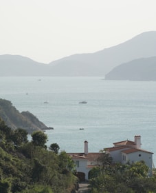 A peaceful coastal scene with a large villa in the foreground, surrounded by lush greenery. In the background, the calm sea stretches out towards a range of gently sloping hills. Several small boats are scattered across the water, contributing to the serene atmosphere.